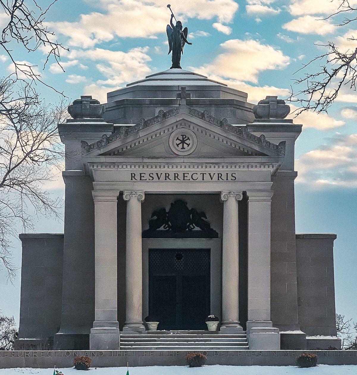 Front of the Bishops' Mausoleum at Mount Carmel Cemetery in Hillside, Illinois: a pale stone building with columns, a triangular pediment inscribed RESVRRECTVRIS and a bronze angel with a trumpet on the roof, against a winter sky