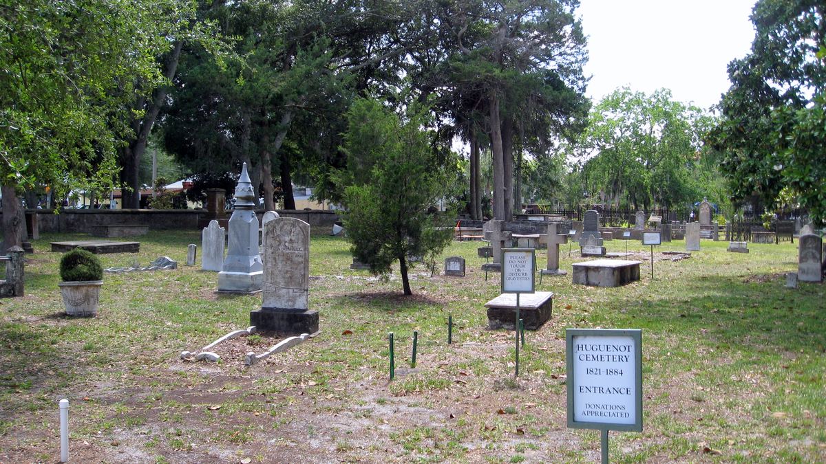 Headstones and monuments under trees inside Huguenot Cemetery in St. Augustine, Florida, with the entrance sign in the foreground